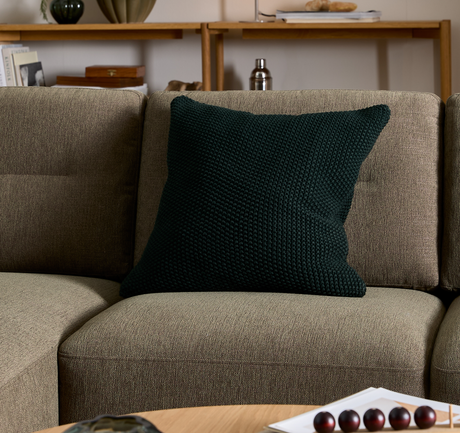 Mörkgrön stickad kudde på beige soffa i vardagsrum med trähylla och dekor i bakgrunden.