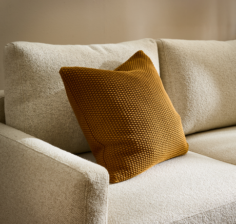 Beige soffa i texturerat tyg med ockragul stickad kudde.
