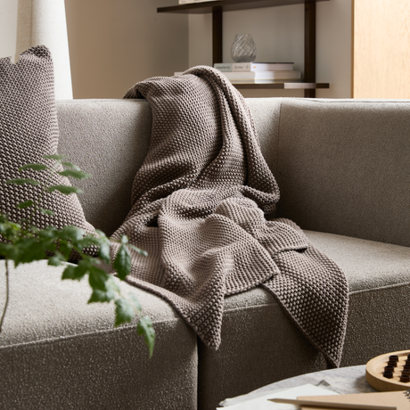 Stickad pläd i taupe ligger över en beige soffa med matchande kudde; växt i förgrunden och böcker samt dekoration på en hylla i bakgrunden.