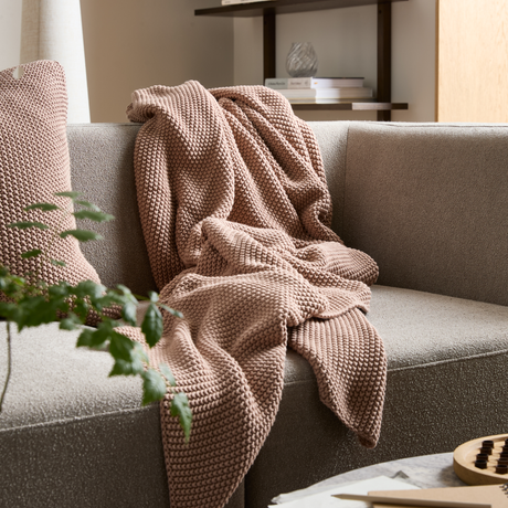 Beige soffa med rosabeige stickad pläd och matchande kudde i ett ljust vardagsrum.