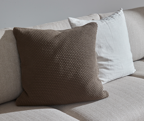 Närbild på beige soffa med stickad brun kudde framför en vit linnkudde i mjukt dagsljus.