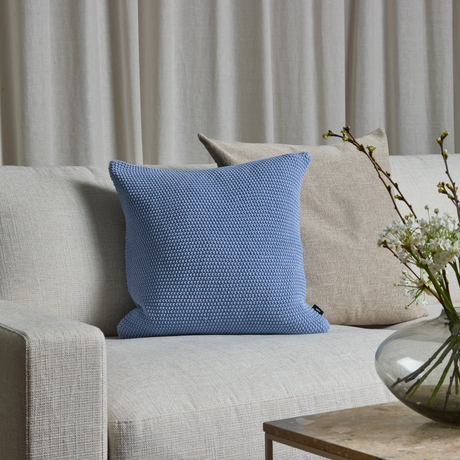 Ljus beige soffa med blå strukturerad kudde framför en beige kudde, glasvas med vita blommor på bord och neutrala gardiner i bakgrunden.