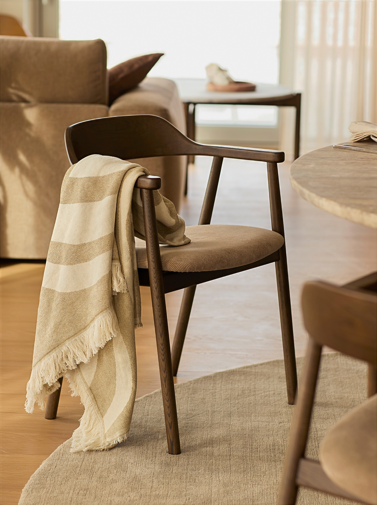 Varm interiör med brun trächair med armstöd och beige pläd över ryglänet vid runt bord, neutral matta och soffa i bakgrunden.