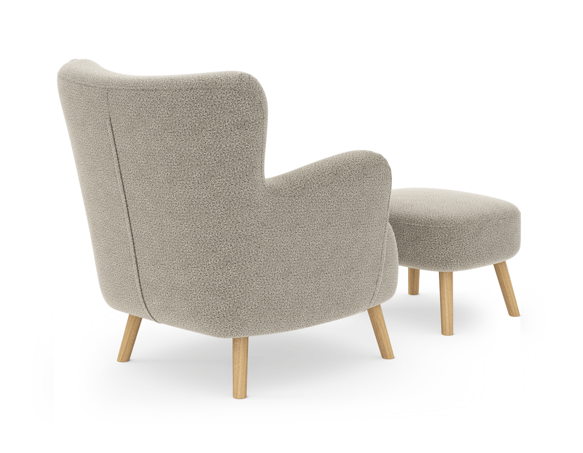 Beige fåtölj med rundad rygg och matchande fotpall på träben, sedd snett bakifrån.