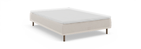 Säng med beige stoppad ram, vit bäddmadrass och träben – sedd snett framifrån mot svart bakgrund.