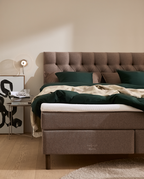 Beige stoppad sänggavel med brun säng och grön bäddning, Nocturne TEMPUR-säng i ett lugnt sovrum med nattduksbord, lampa, böcker och grafisk tavla.