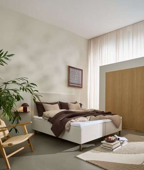 Ljust, skandinaviskt sovrum med beige kontinentalsäng, bruna och sandfärgade sängkläder, väggmonterad lampa, stor grön växt, takhöga ljusa gardiner, trägarderob och mjuk matta; böcker och bricka ligger vid fotänden.
