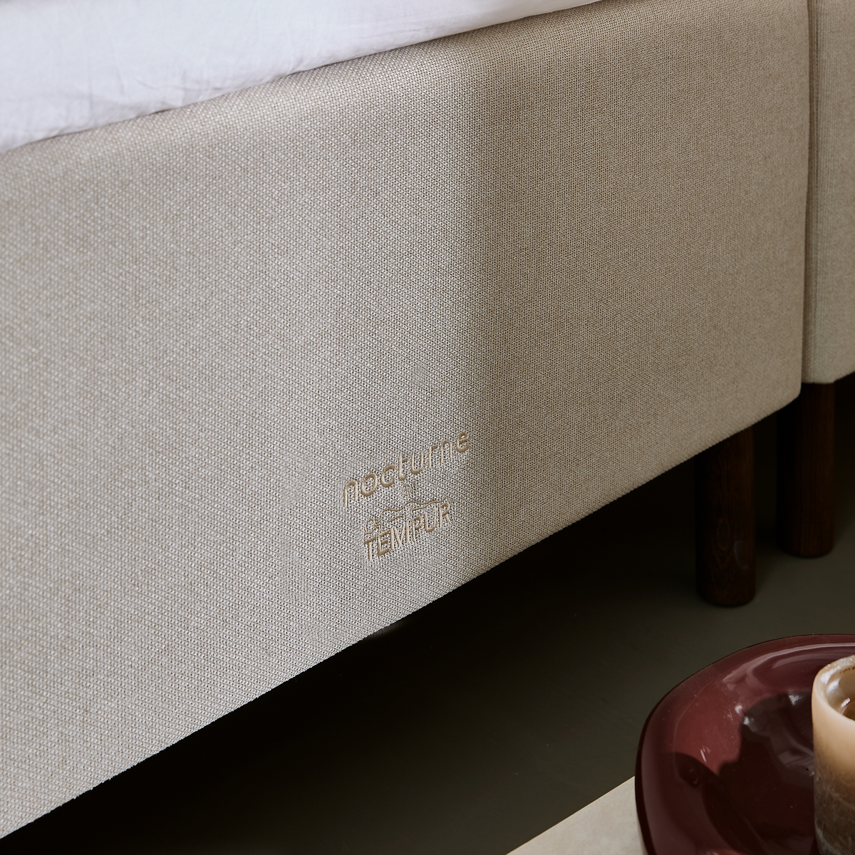 Närbild av beige sängbas med broderad logga 'nocturne by TEMPUR', träben samt ett ljus på ett mörkrött fat i hörnet.
