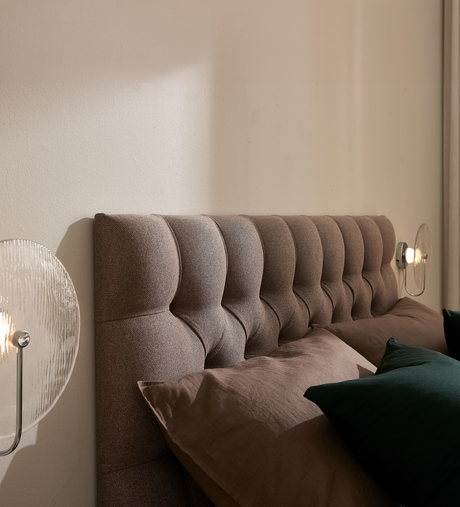 Närbild av en polstrad, knapp-tuftad sänggavel i beige med bruna och mörkgröna kuddar samt glasvägglampor i ett lugnt sovrum.