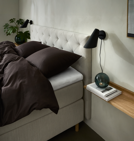 Sovrumshörn med beige säng och knapptuftad sänggavel, mörkbrunt bäddset, svart vägglampa och smal trähylla med böcker och grön glaslykta; grön växt bredvid.