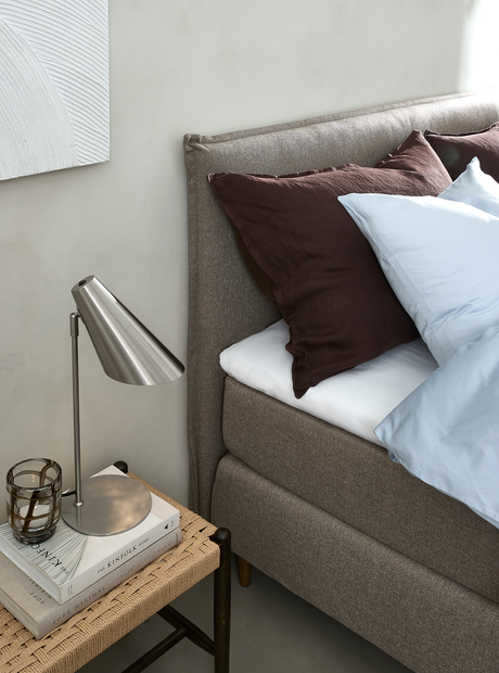 Närbild av ett sänghörn med grå stoppad gavel, bruna och ljusblå kuddar samt ett nattduksbord med stålfärgad bordslampa, glaslykta och böckerna The Kinfolk Home och Soft Minimal.