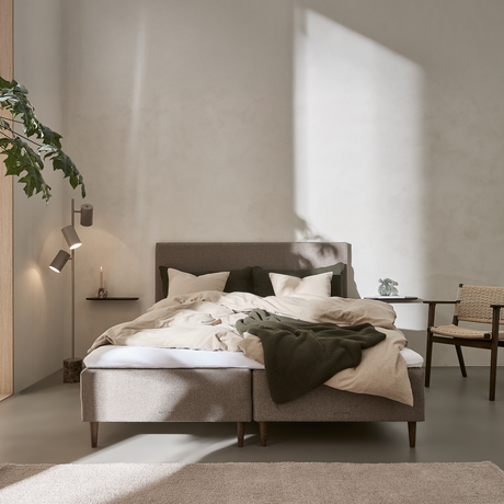Ljust sovrum med solstrålar på vägg, grå stoppad säng med beige bäddning och grön pläd, växt till vänster, litet sidobord med ljus och stol av rotting.