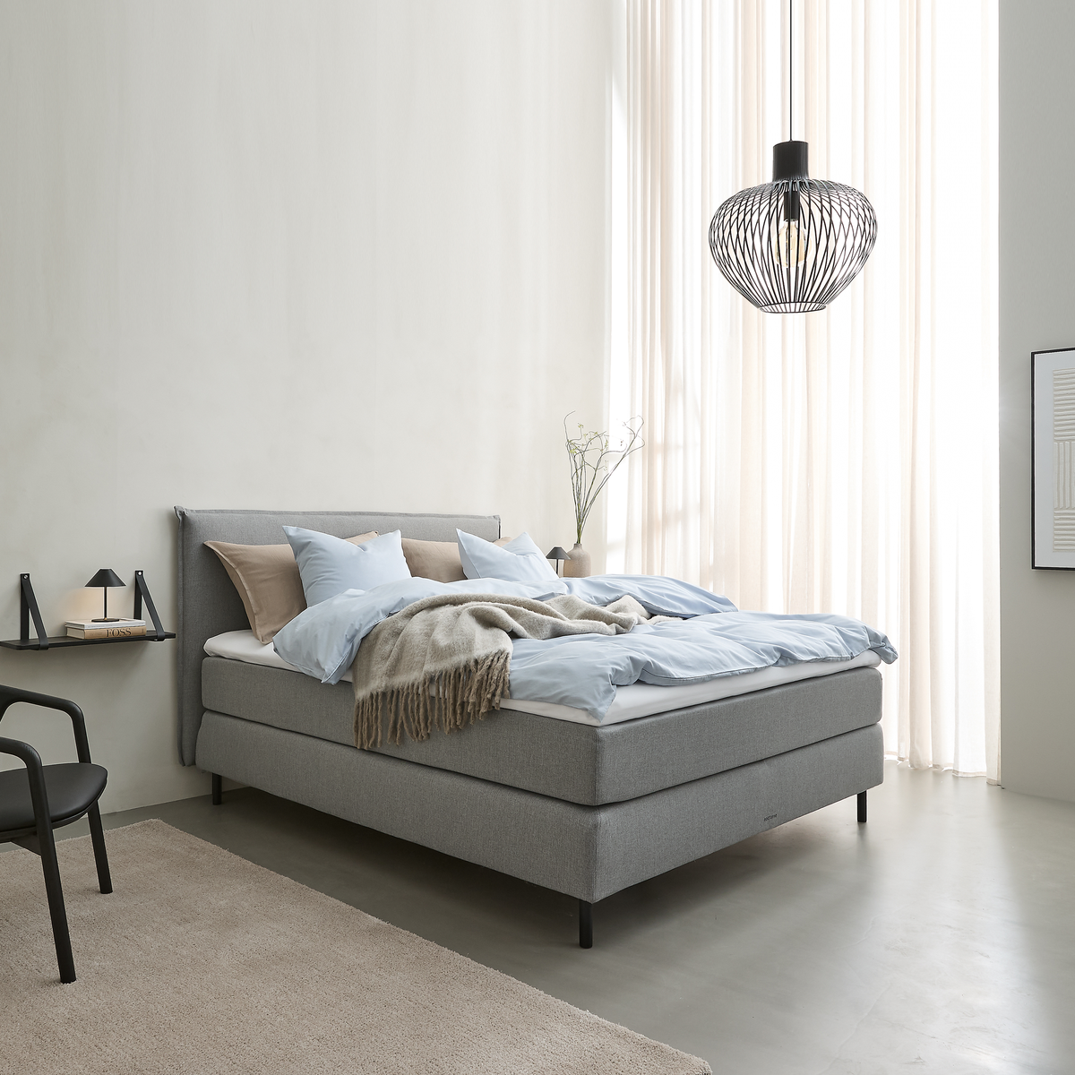 Ljust sovrum i skandinavisk stil med grå klädd säng, ljusblått bäddset och beige pläd, svart trådpendel och tunna gardiner som släpper in dagsljus.