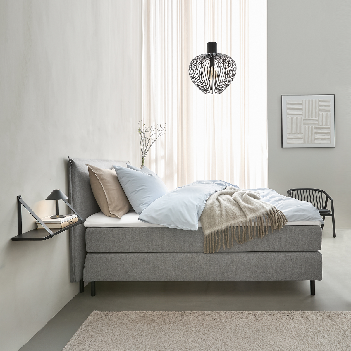 Minimalistiskt sovrum med grå säng, ljusblått bäddset, beige pläd, svart pendellampa och liten vägghylla med lampa och böcker; mjukt dagsljus genom skira gardiner.