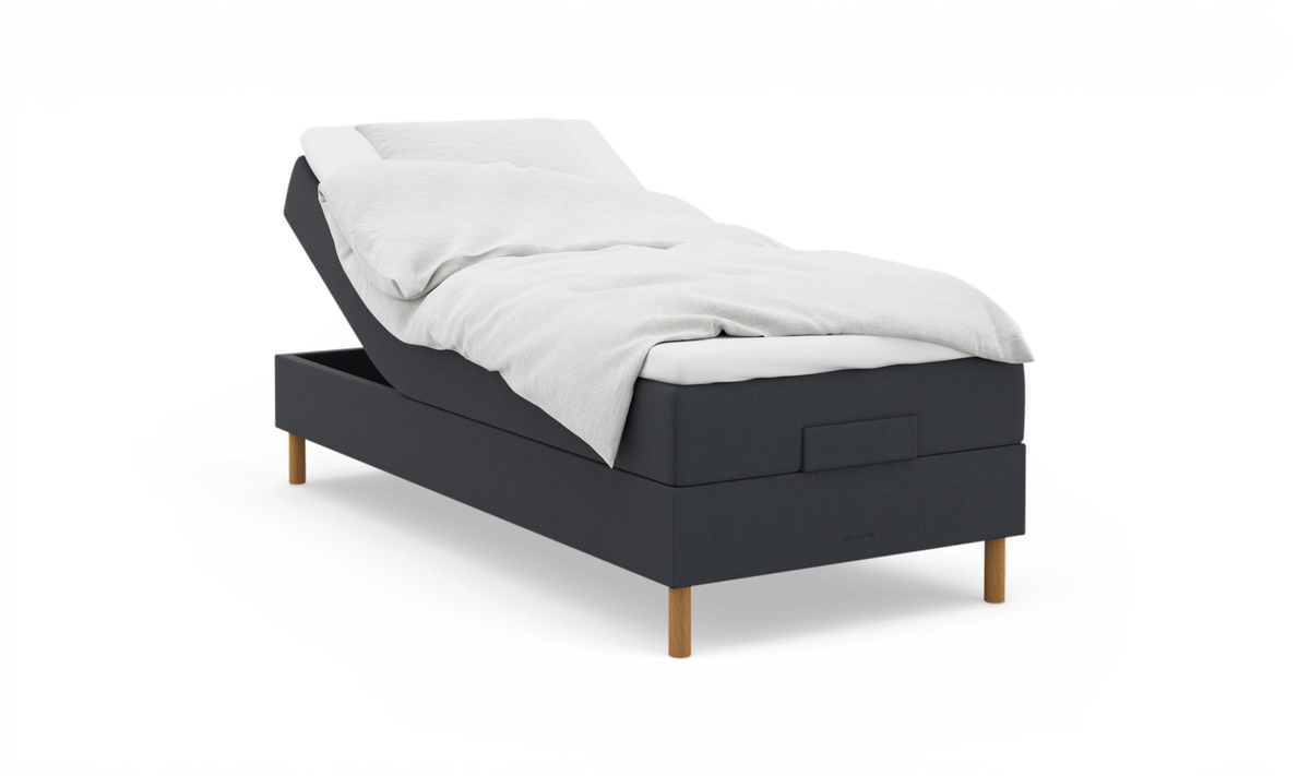 Justerbar säng i mörkgrå textil med vita sängkläder och träben; ”nocturne” syns på sängkanten.