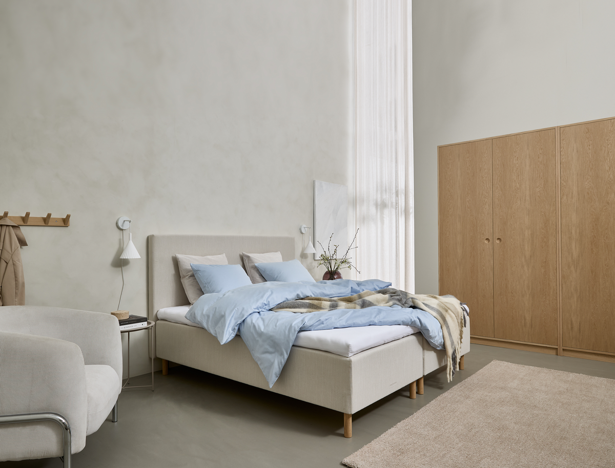 Ljust, minimalistiskt sovrum med beige stoppad säng, ljusblått bäddset, vägglampetter, trägarderob och mjuk matta vid ett högt fönster med skir gardin.