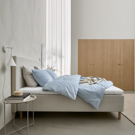 Ljust, minimalistiskt sovrum med beige säng, ljusblått påslakan och kuddar, vägglampa, litet sidobord med böcker och garderober i ljus ek.