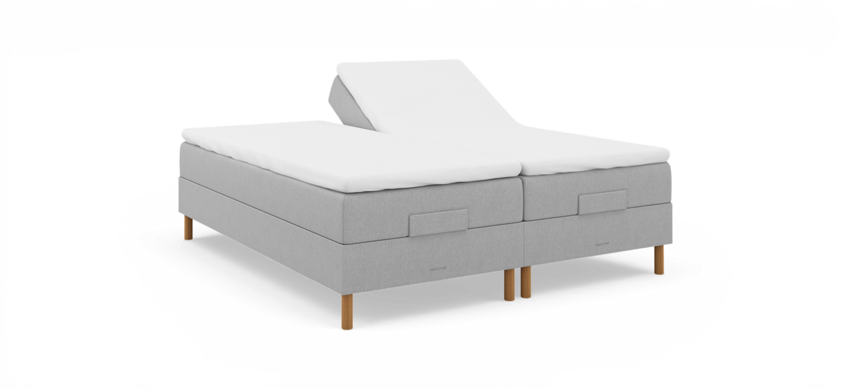 Justerbar dubbelsäng i ljusgrå textil med vit bäddmadrass, träben och logotypen nocturne; ena huvudänden är upphöjd.