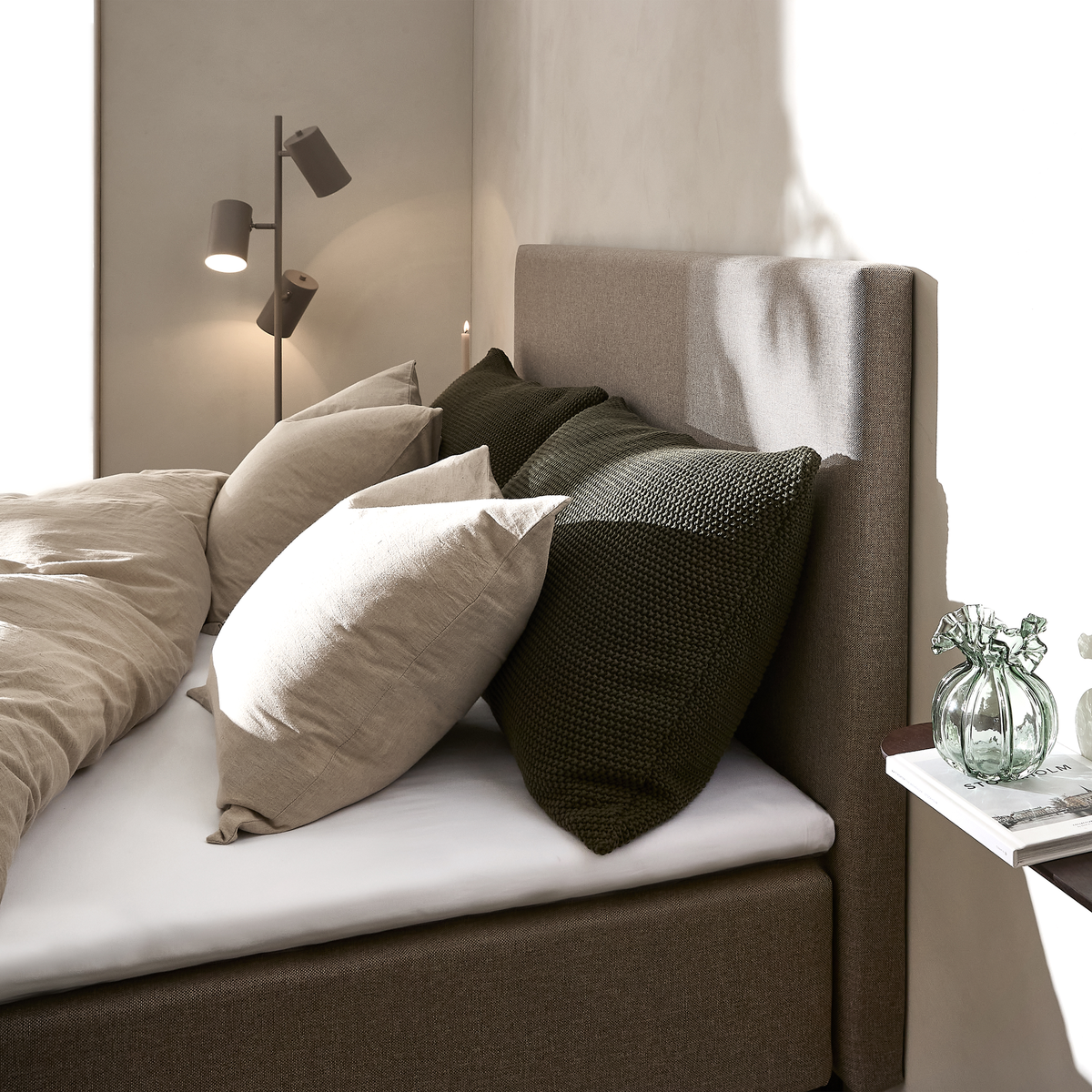 Närbild av en bäddad säng med beige och mörkgröna kuddar, stoppad brun sänggavel, en golvlampa samt en liten hylla med glasvas och bok i mjukt dagsljus.