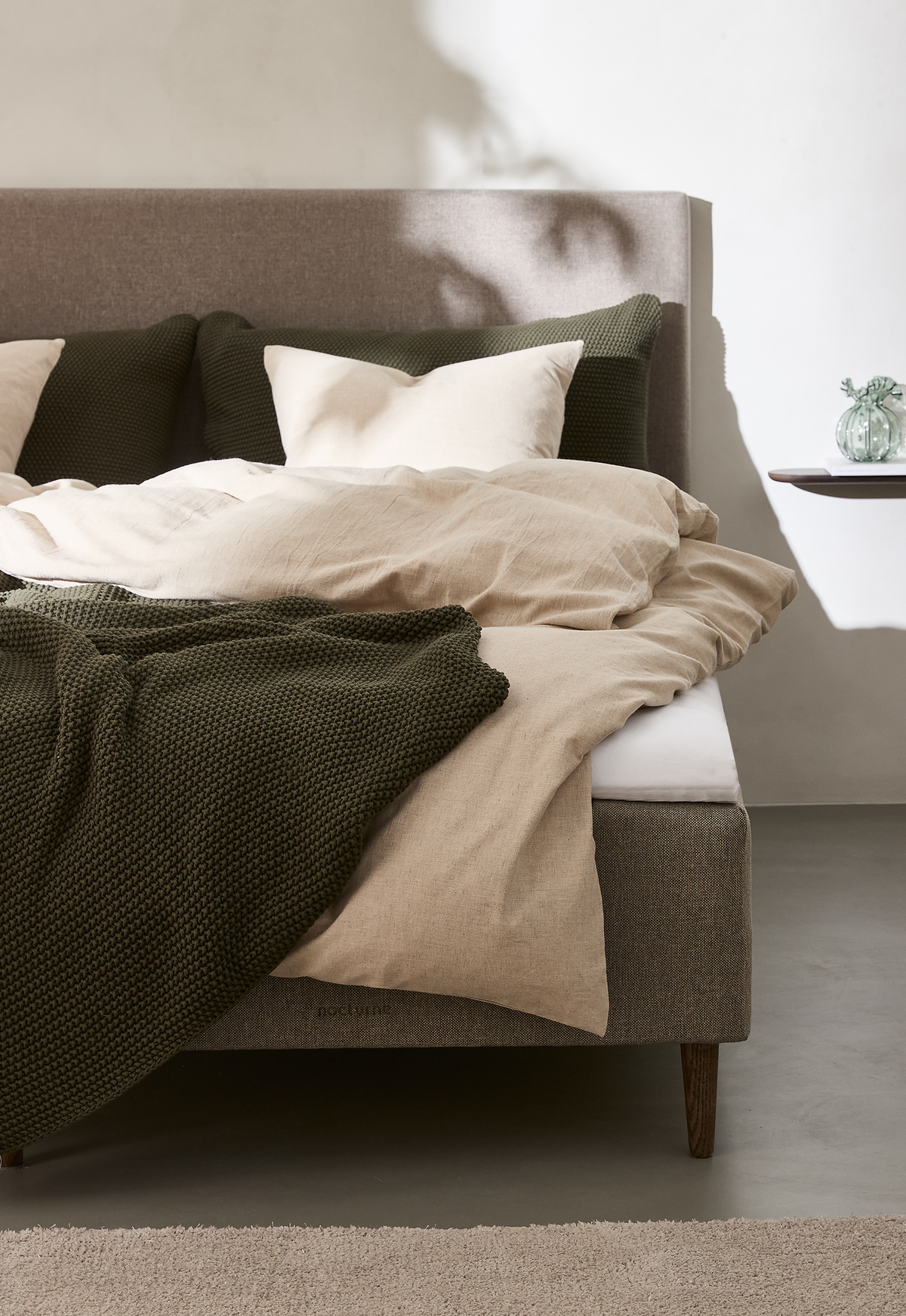Beige linnesängkläder med mörkgrön stickad pläd på en stoppad säng i ett ljust, minimalistiskt sovrum.