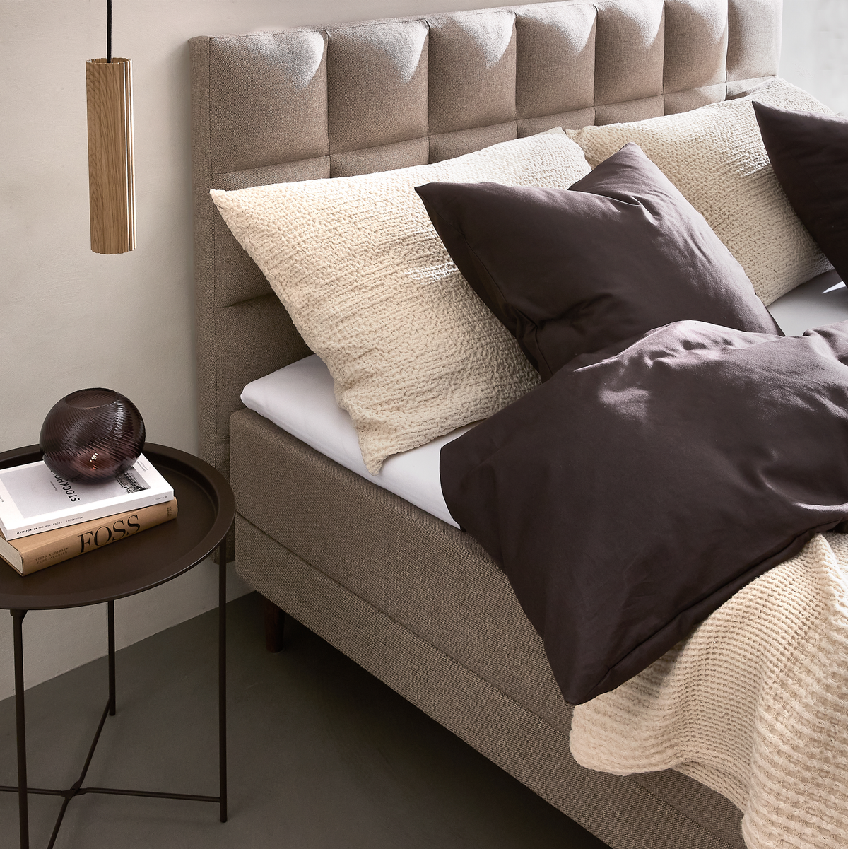 Beige stoppad säng med mörkbruna och krämfärgade kuddar, stickad pläd, runt sängbord med bok med titeln FOSS och en rökfärgad glasdekoration samt hängande träpendel.