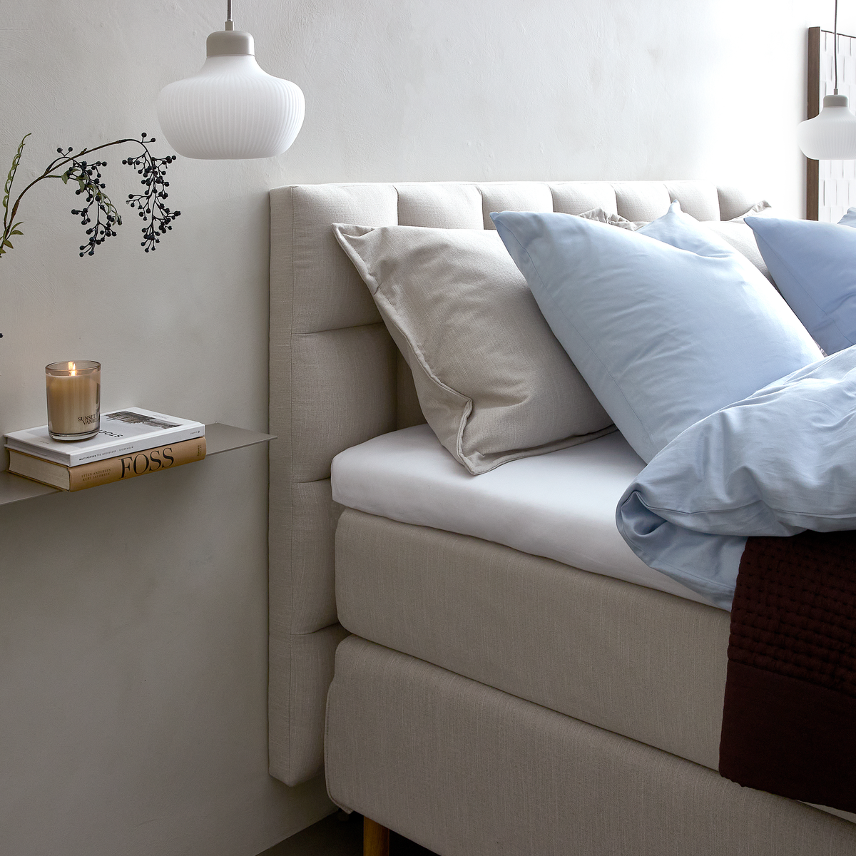 Ljus och rofylld sovrumsdetalj med beige kontinentalsäng, ljusblått påslakan, kuddar, en mörk stickad pläd och en vit pendellampa; på en väggmonterad hylla står ett ljus och böcker, varav en med titeln FOSS.