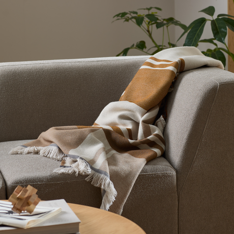 Beige soffa med en mönstrad pläd i vitt och bruna toner med fransar, växt i bakgrunden och soffbord med böcker i förgrunden.