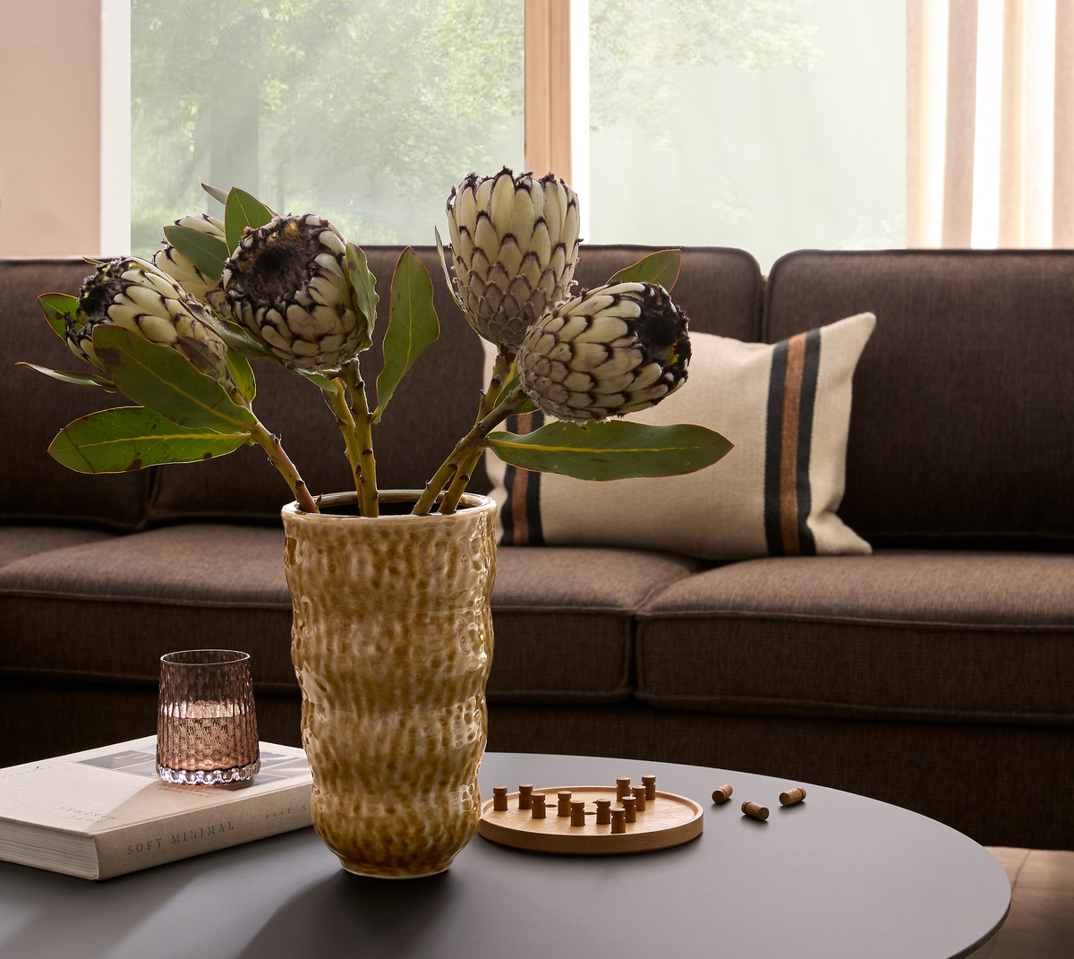 Proteablommor i en strukturerad beige vas på ett soffbord med glas, boken Soft Minimal och ett litet träspel; brun soffa med randig kudde och fönsterljus i bakgrunden.