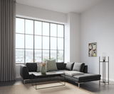 Ljus skandinavisk vardagsrumsscen med grå hörnsoffa, slankt soffbord i svart metall, vas med torkade blommor, stor fönstervägg med stadsutsikt, abstrakt tavla och glas-golvlampa; beige matta och pläd på schäslongen.