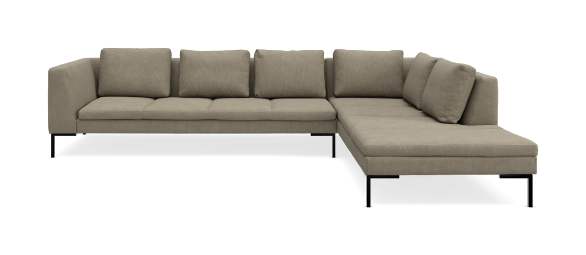 Beige L‑soffa med schäslong till höger, smala svarta ben och lösa ryggkuddar.
