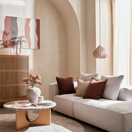 Ljust skandinaviskt vardagsrum med beige soffa, roströda och sandfärgade kuddar, runt trä/sten-soffbord med vas, ljus och boken CHLOE SEVIGNY; ribbad skänk, abstrakt väggkonst och rosa pendellampa i mjukt dagsljus vid skira gardiner.