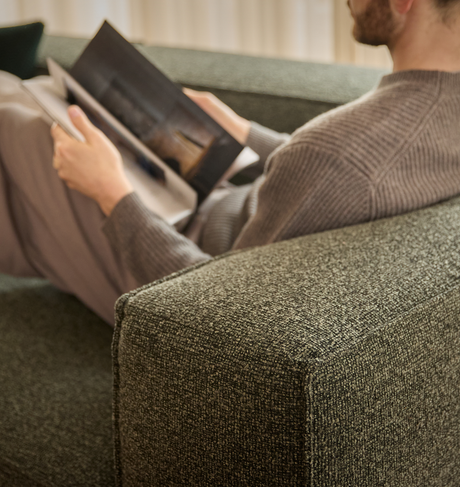 Person sitter avslappnat på en grön textilsoffa och bläddrar i en tidskrift.