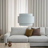 Ljus, minimalistisk vardagsrumsscen med ljusgrå soffa, två kuddar i brunt och oliv, stor vit pendellampa, ljus stenbord med ljuslykta och liten glasvas samt beige gardiner i bakgrunden.