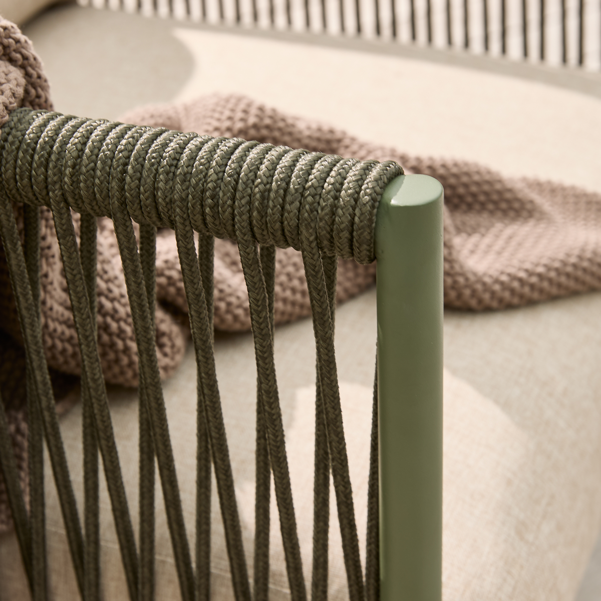 Närbild av grön utestol med flätad repdetalj, beige dyna och stickad brunbeige pläd.