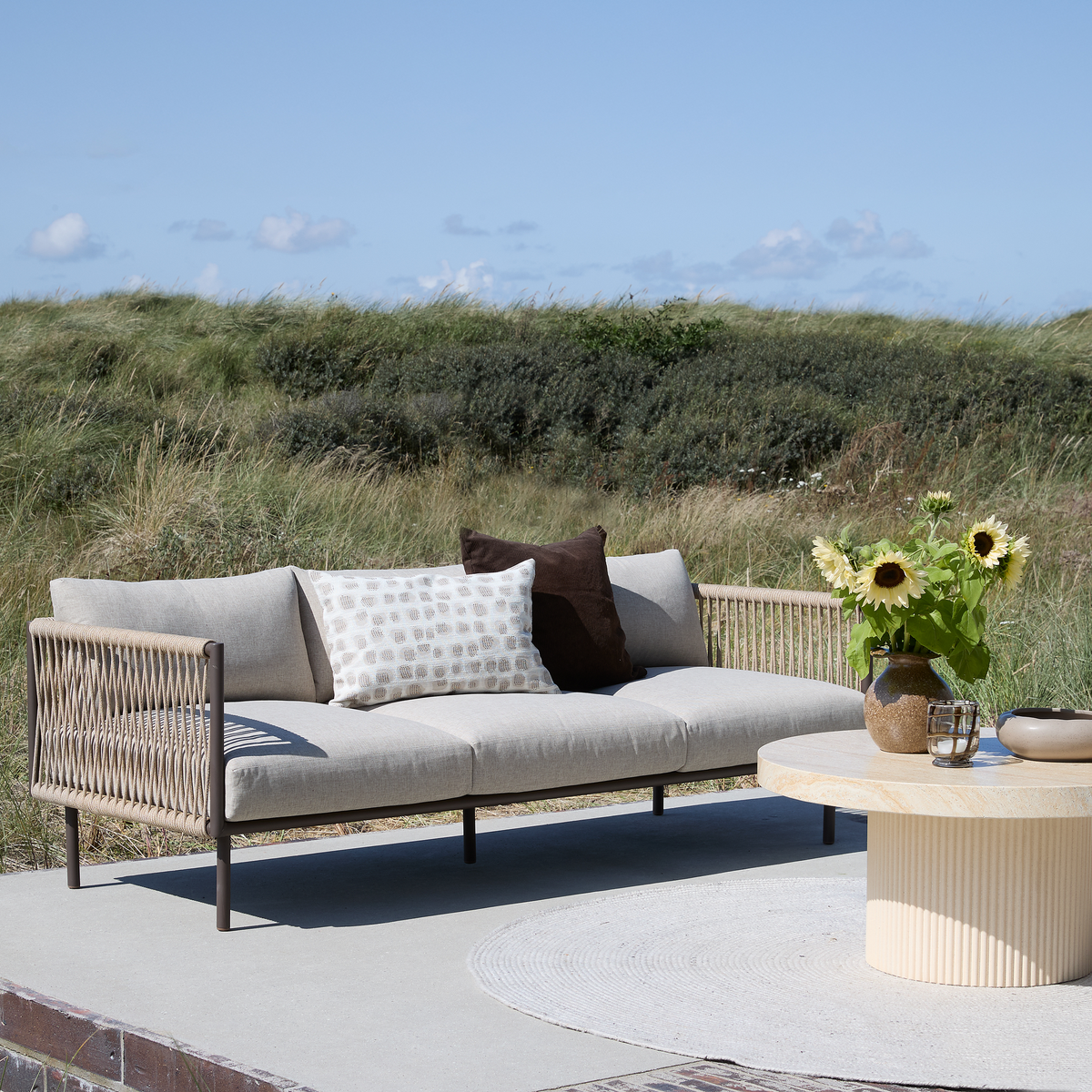 Utomhusmiljö med beige flätad loungesoffa med kuddar och runt ljust stenbord med vas med solrosor på en terrass, med gräsbevuxna dyner och blå himmel i bakgrunden.