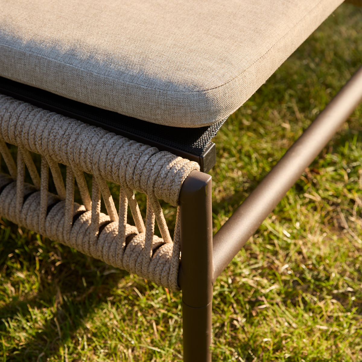 Närbild av utomhuslounge-stol med beige dyna, flätad repdetalj och brun metallram på gräsmatta i solsken.