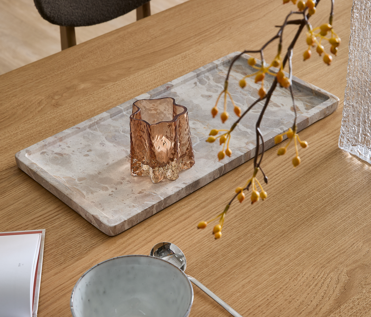 Träbord med rektangulärt marmorfat, bärnstensfärgad glasvas och en gren med gula bär; skål och sked i förgrunden.
