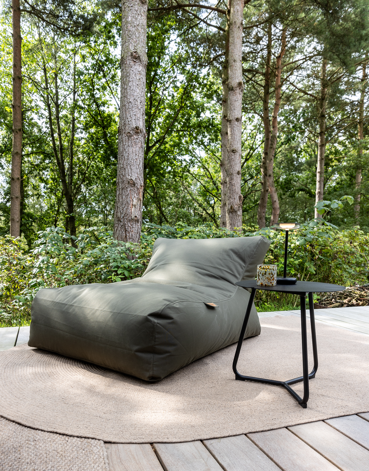 Grön sittsäck på träterrass med rund beige matta och liten svart sidobord med lampa och kopp, omgiven av skog i dagsljus.