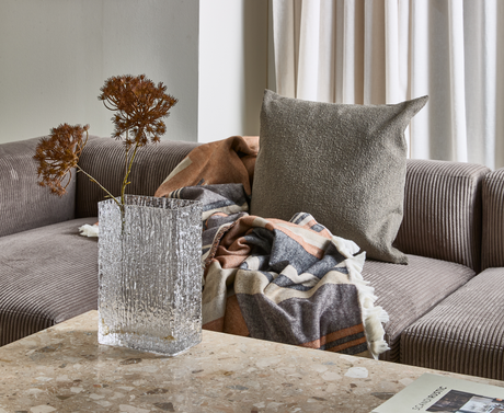 Soffhörn med grå ribbad soffa, beige kudde och mönstrad pläd; glasvas med torkade kvistar på ett terrazzo-bord samt ett magasin med titeln Scandi Rustic i hörnet.