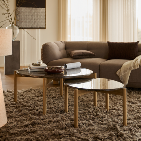 Vardagsrum med två runda soffbord i mörk sten och ek, på brunt ryamatta framför beige modulsoffa med kuddar och pläd, mjuka gardiner och varm belysning.