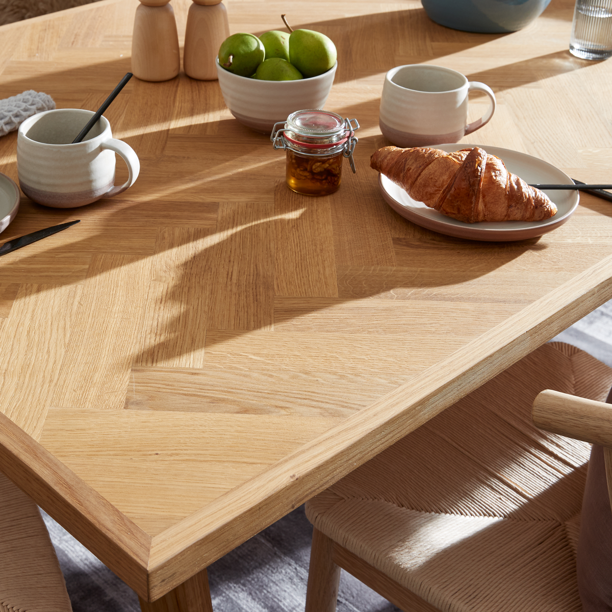 Närbild av ett ljust ekbord med frukost – croissant på fat, två keramikmuggar, skål med gröna äpplen och liten burk honung i soligt dagsljus.