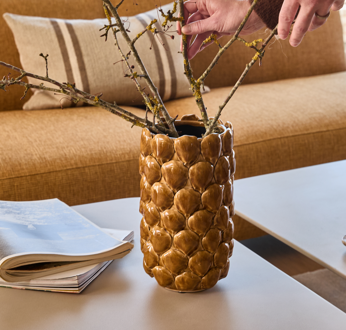 Hand som arrangerar kvistar i en brun, strukturerad keramikvas på soffbord; tidning och beige soffa med kudde i bakgrunden.
