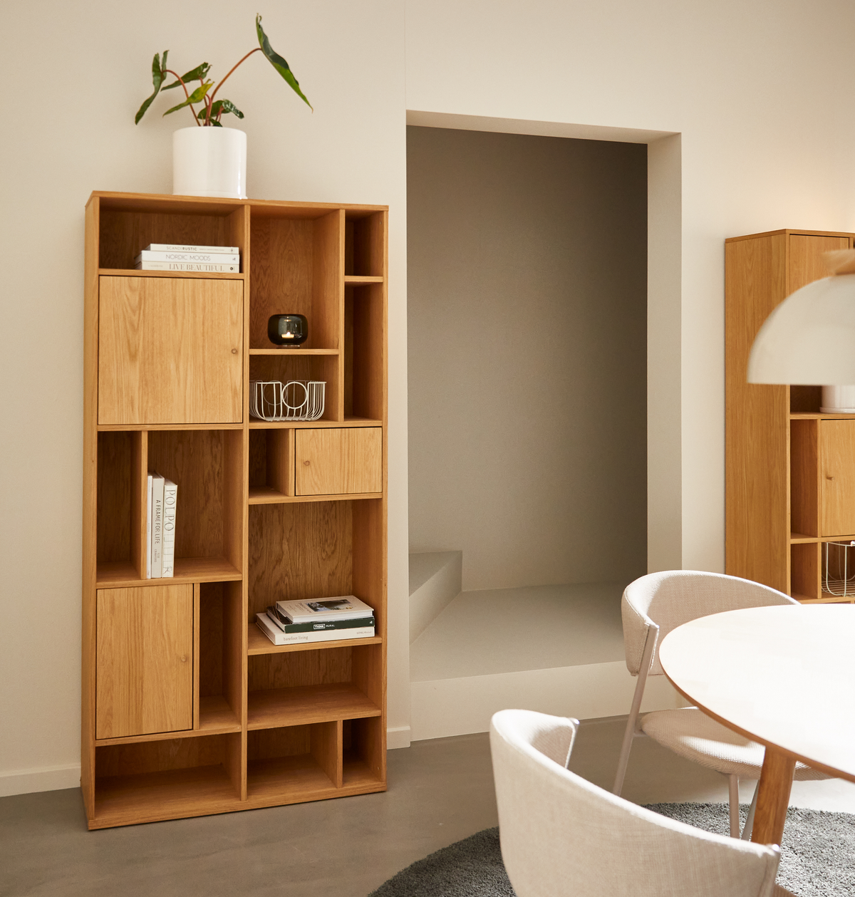 Ljus skandinavisk matplats med ekhylla i moduldesign; på hyllan ligger böcker med titlarna “NORDIC MOODS” och “NORM ARCHITECTS”, en vit kruka med grön växt och en vit trådkorg; runt bord med stoppade stolar och vit pendellampa i neutrala toner.