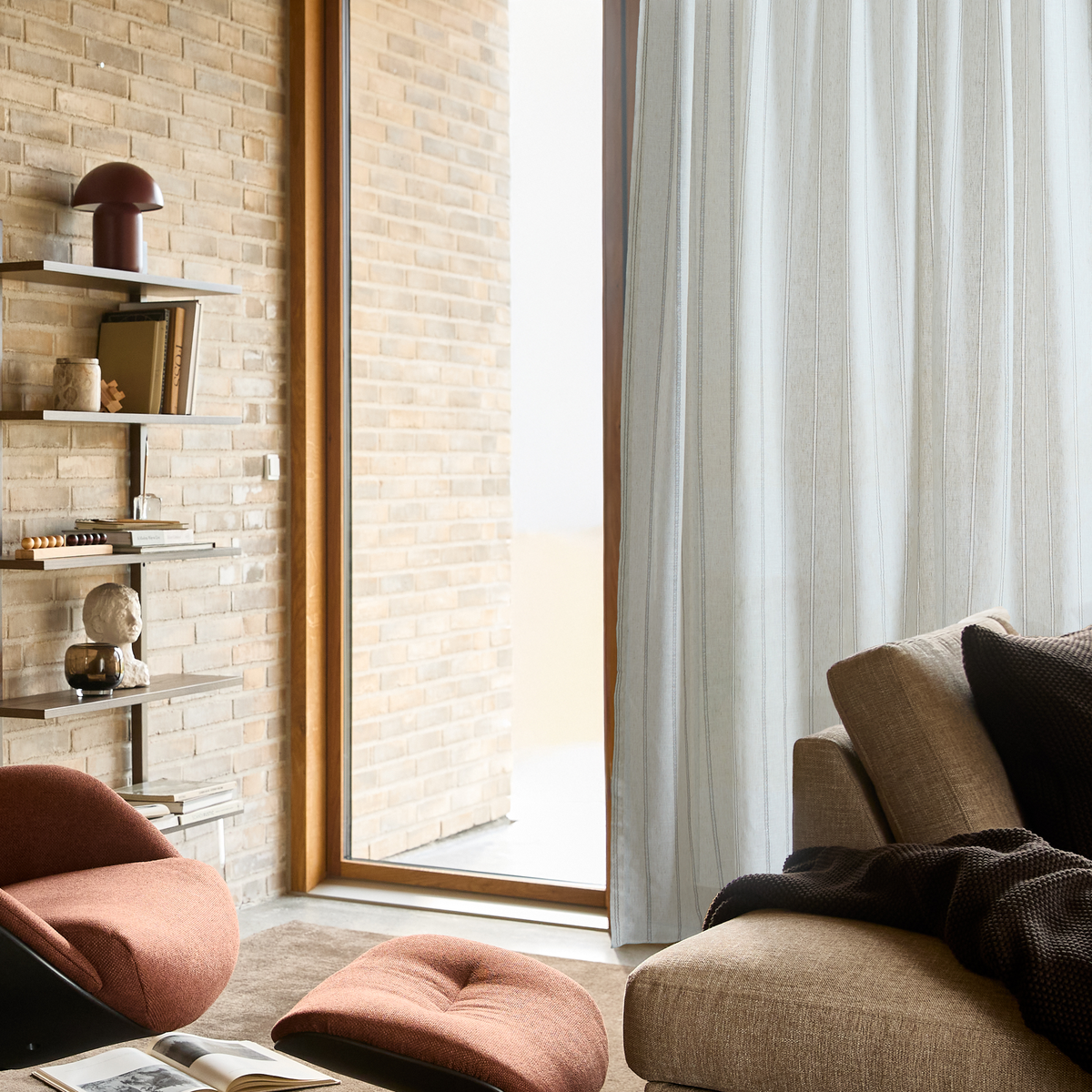 Ljus vardagsrumshörna med beige soffa med mörk pläd och kuddar, skir gardin vid stort fönster, väggmonterade hyllor med böcker, liten bordslampa och skulptur samt rostfärgad fotpall på matta.