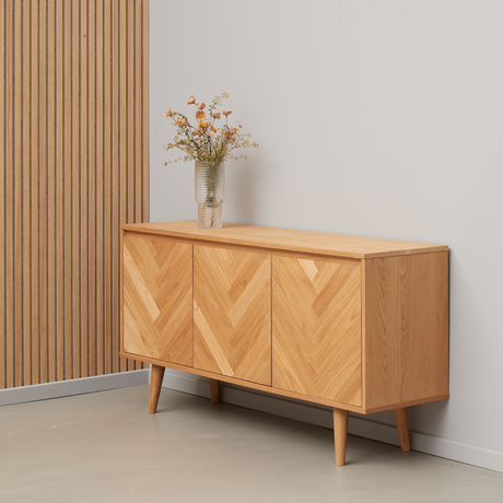 Ljust sideboard i ek med fiskbensmönstrade dörrar och koniska ben, en vas med vilda blommor ovanpå mot grå vägg och ribbad träpanel.