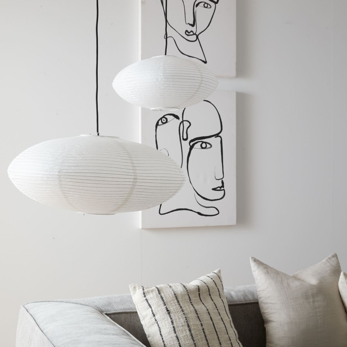 Två vita rispendlar hänger över en grå soffa med neutrala kuddar; bakom syns två svartvita lineart-tavlor med ansikten.