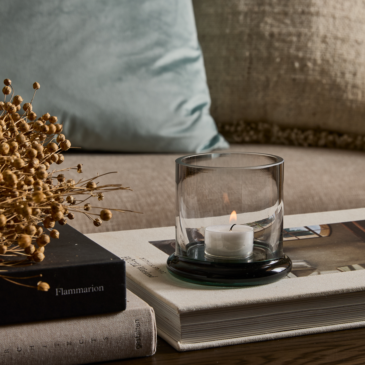 Glaslykta med tänd värmeljus på staplade designböcker, bland annat Flammarion, med torkade kvistar på ett soffbord i vardagsrum med mjuka kuddar.