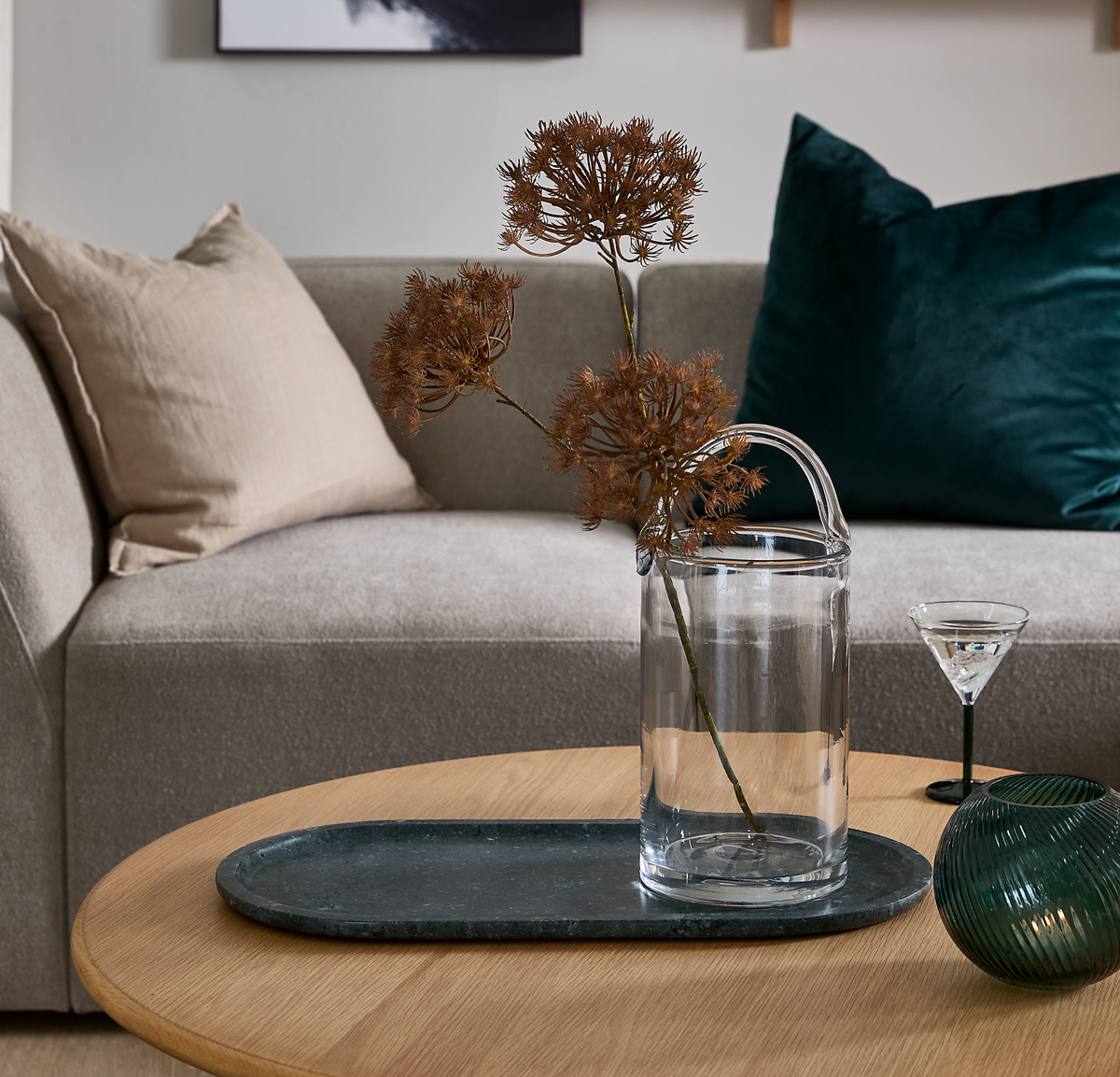 Beige soffa med kuddar bakom ett runt soffbord i trä med mörk stenbricka, glasvas med torkade kvistar och ett litet cocktailglas.