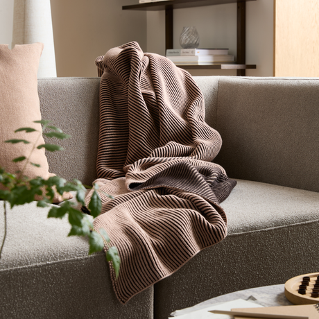 Randig stickad pläd i beige och mörkbrunt ligger över en ljusgrå soffa med kudde; växt och hylla med böcker i bakgrunden.
