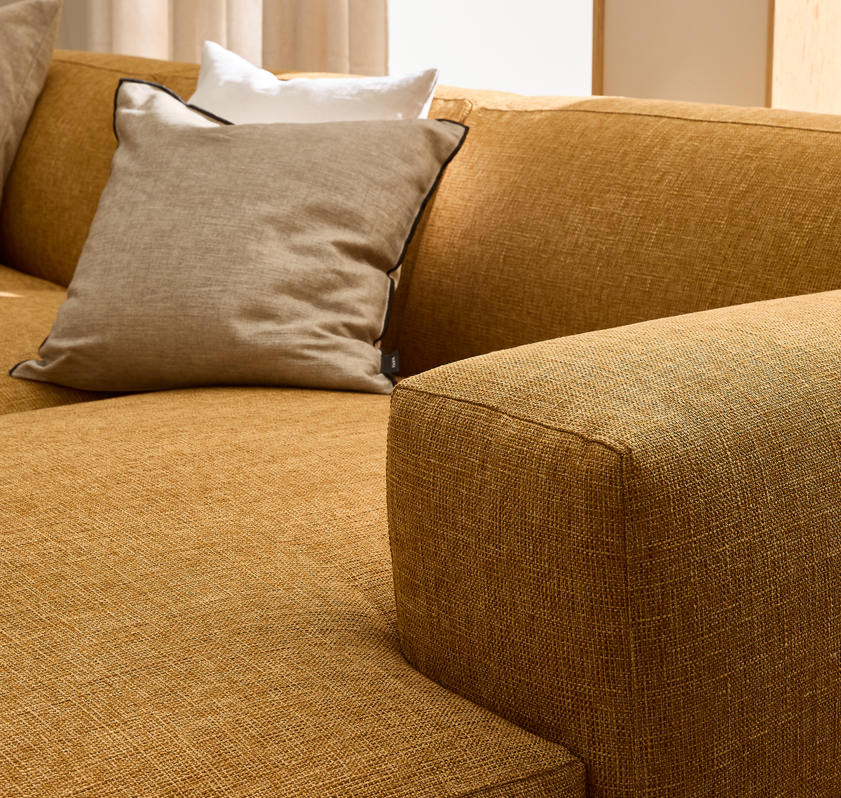 Närbild av en senapsgul tygsoffa med en beige kudde med mörk kant och en vit kudde bakom i ett ljust vardagsrum.